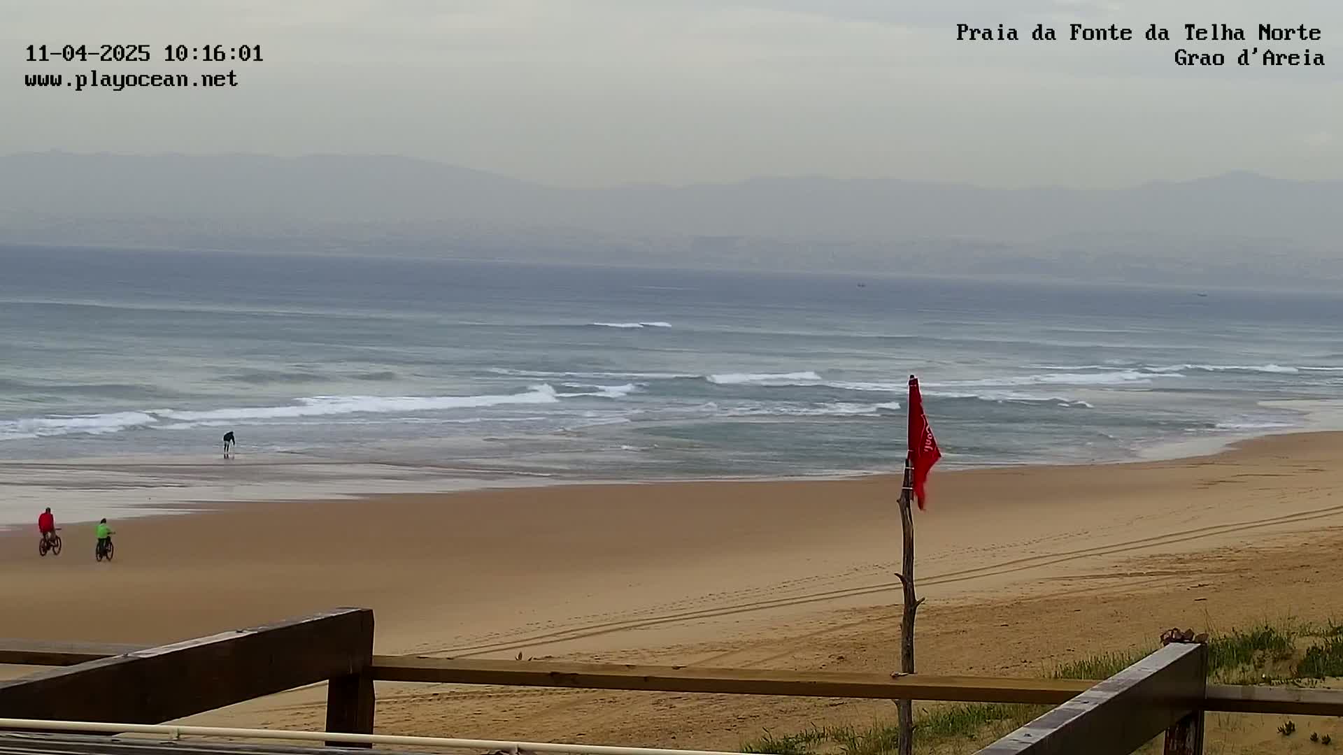 A hazy beach scene shows a sandy shore with a few people riding bicycles, small waves breaking in the ocean, and a red flag on a post in the foreground.