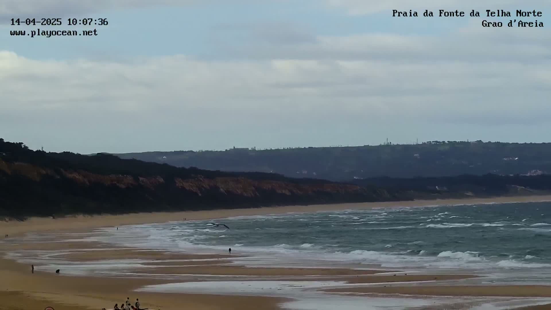 A wide sandy beach with moderate waves under a cloudy sky stretches to a dark cliff in the background.