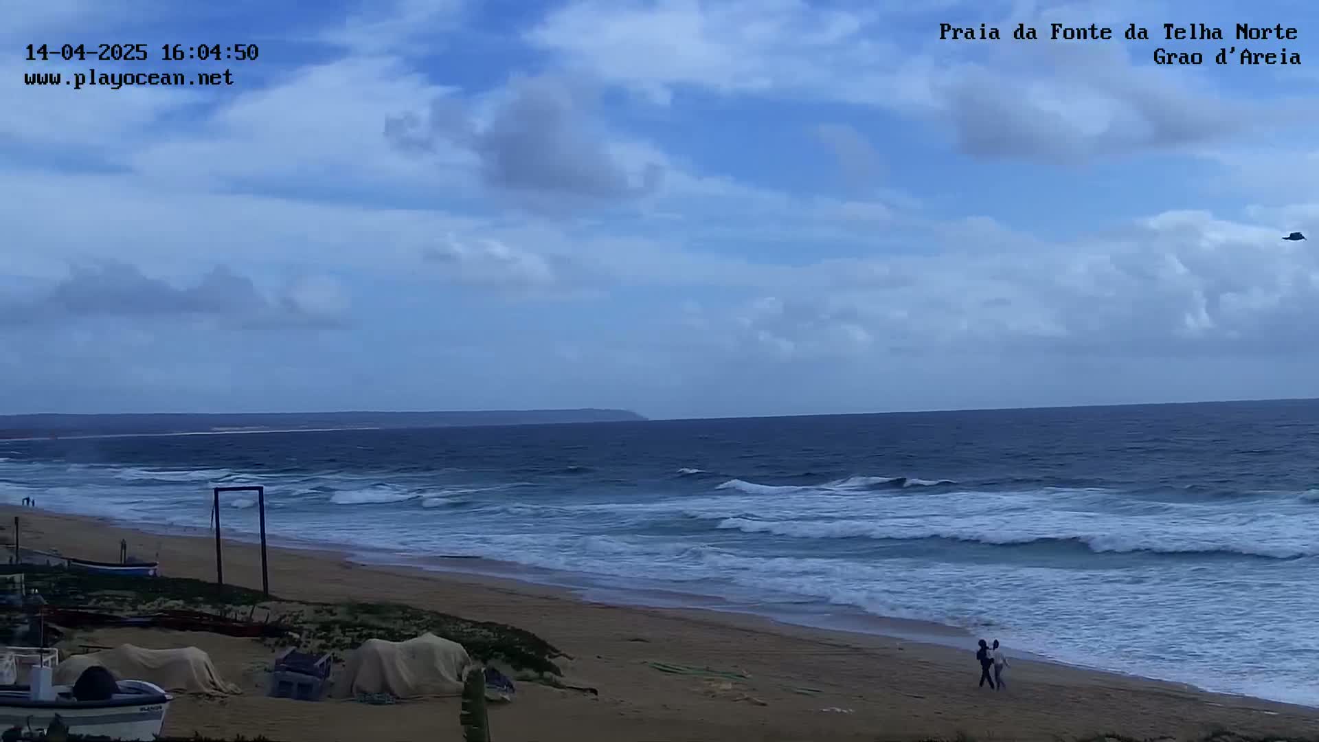 A sandy beach with a calm ocean under a partly cloudy sky, with a couple walking along the shoreline.