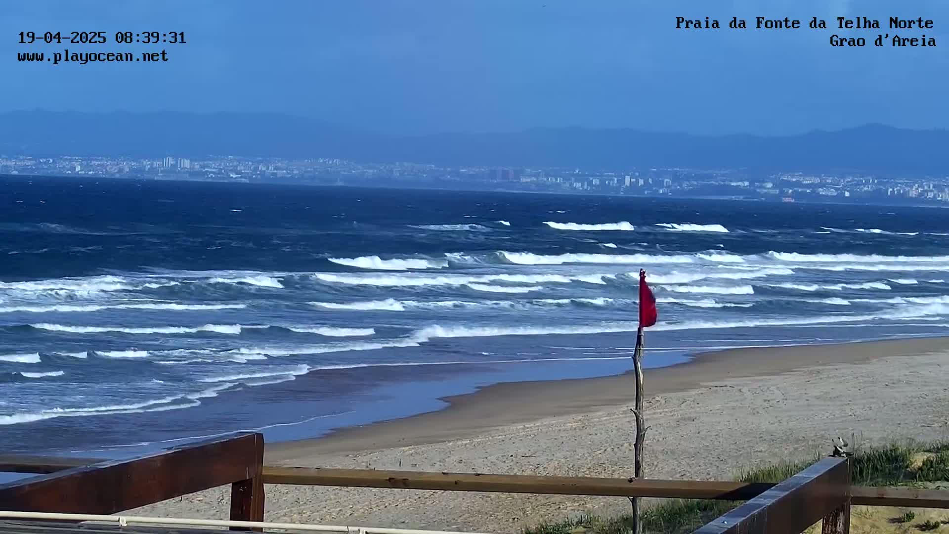 A sandy beach with a red flag, moderate waves rolling in from a blue ocean under a clear sky, and a distant cityscape visible on the horizon.