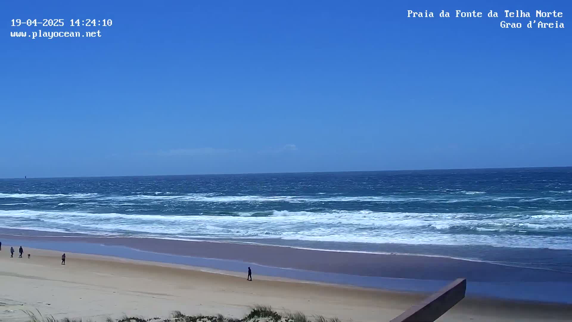 A sandy beach with a few people walking on it, under a clear blue sky, and a choppy ocean with whitecaps.