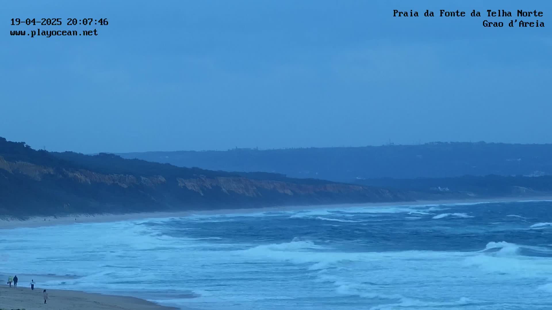 Under a clear blue sky, a sandy beach meets a restless ocean with moderate waves rolling towards the shore, where a few people are walking.