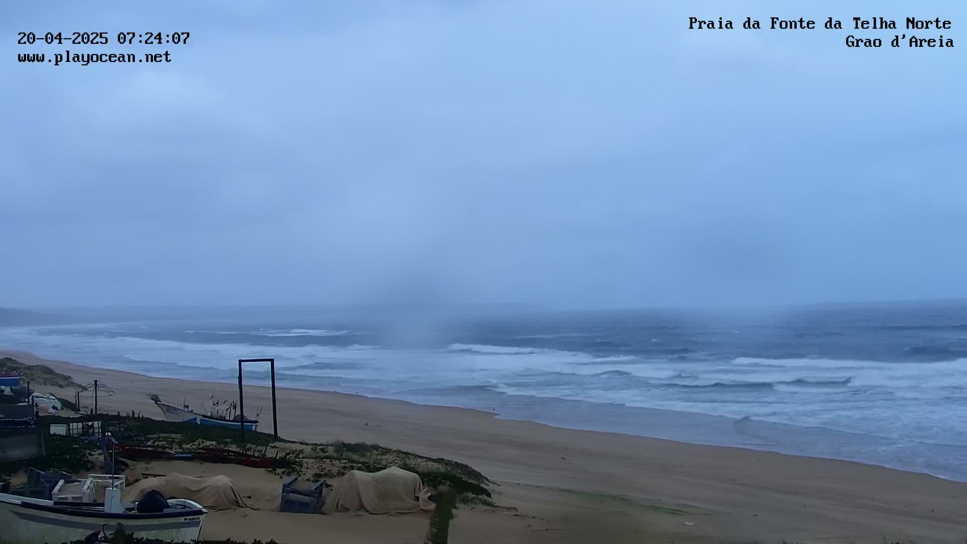 A wide sandy beach with choppy waves under an overcast sky is shown, with several boats and covered items visible on the shore.