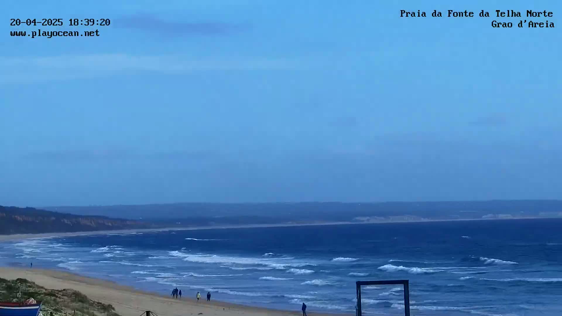 A mostly clear, blue sky overlooks a sandy beach with moderate waves and a few people walking along the shore.
