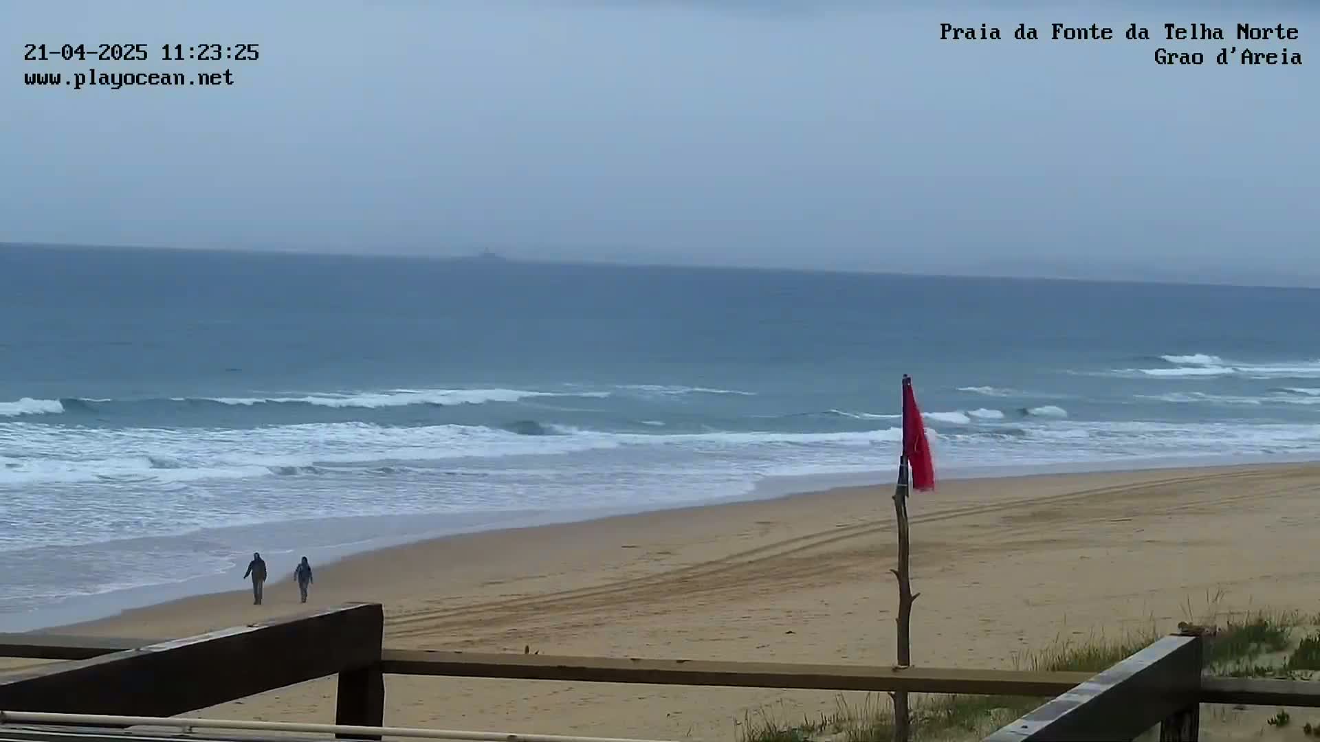 Two people walk along a sandy beach with moderate waves under an overcast sky.
