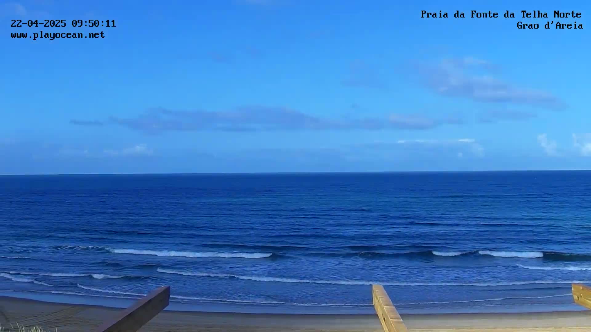 A mostly sunny day shows a wide expanse of dark-blue ocean with small waves lapping a sandy beach, viewed from a slightly elevated vantage point.
