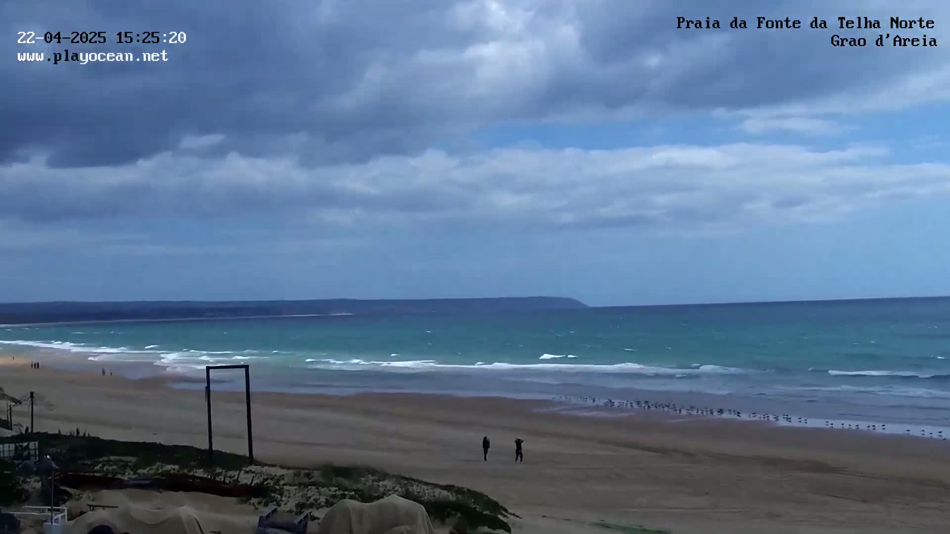 A wide sandy beach stretches to a teal ocean with small waves under a mostly cloudy sky.