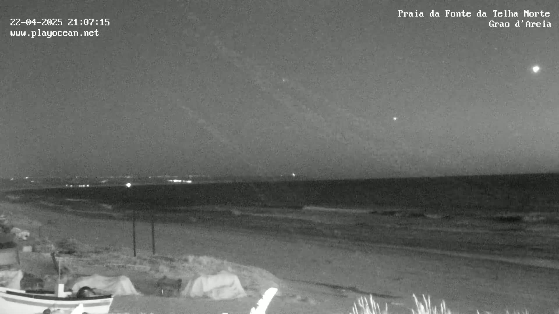 A dark, nighttime beach scene shows a calm ocean with small waves lapping the shore under a clear sky with a few visible stars.