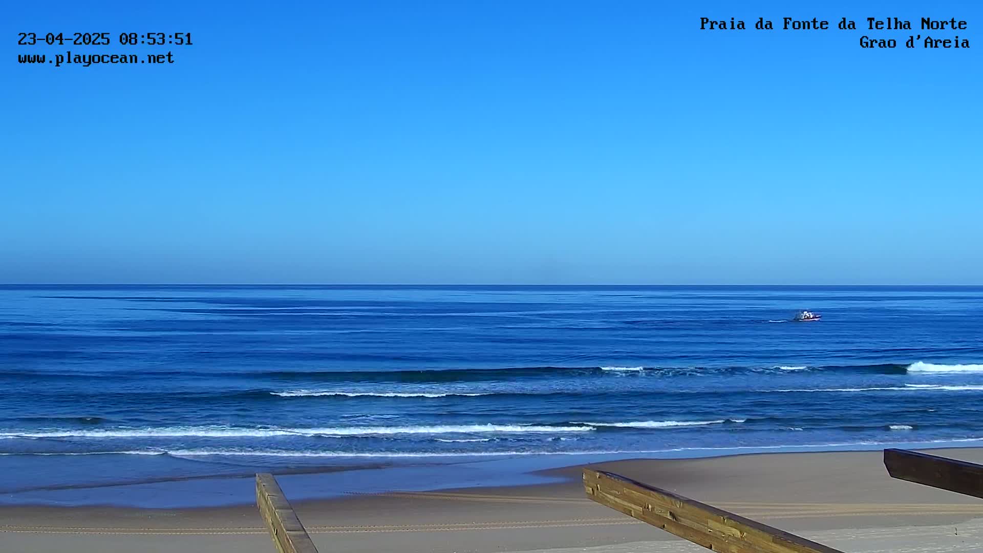 A small boat is visible on a calm, blue ocean under a clear blue sky, with a sandy beach and wooden structures in the foreground.