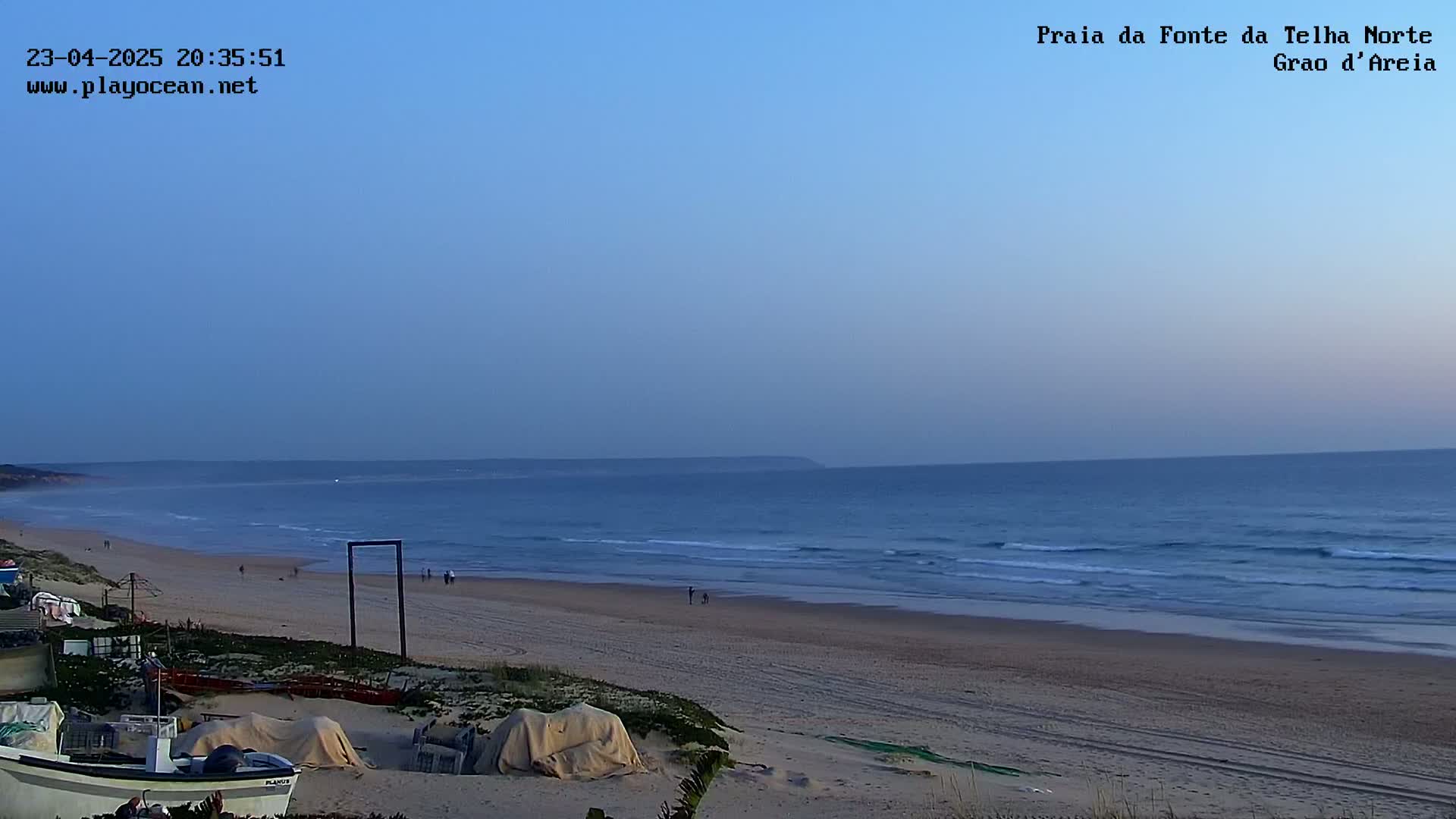 A sandy beach with gentle waves under a twilight sky, a few people are visible in the distance.
