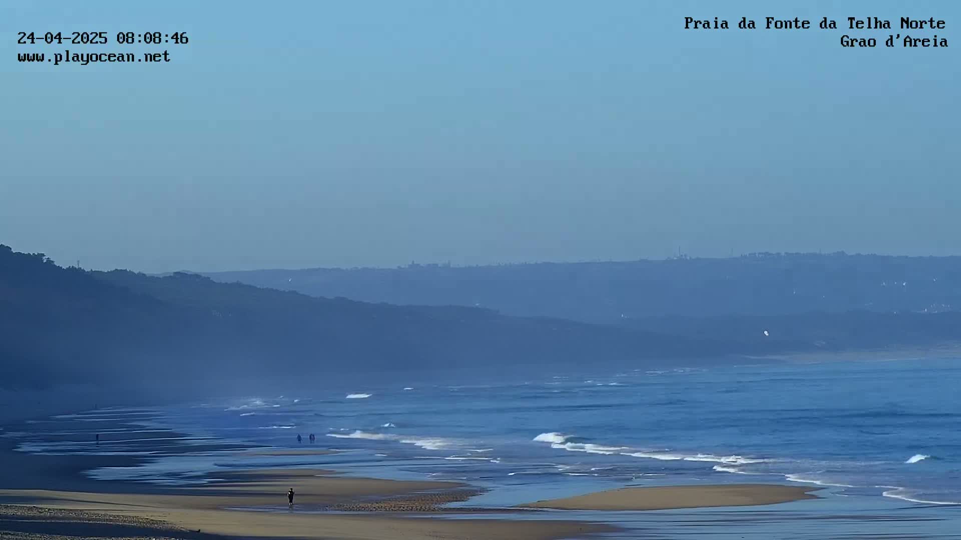 A hazy, clear day reveals a long, sandy beach with gentle waves lapping the shore, a few people walking on the beach, and a distant coastline under a pale blue sky.