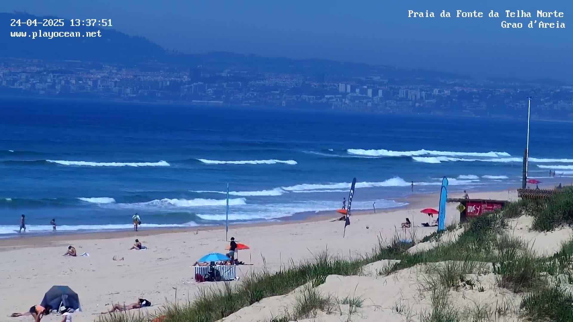 A sandy beach with scattered people, beach umbrellas, and surfboards, features gentle waves lapping the shore under a clear, sunny sky, with a distant city visible across the water.