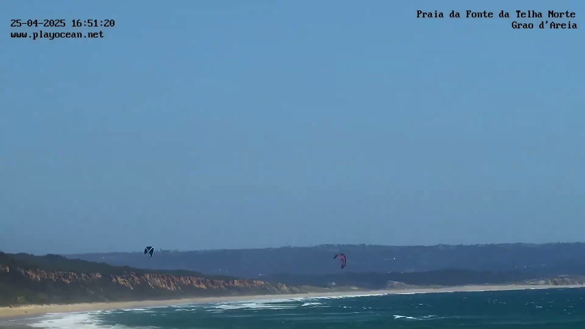 Two kitesurfers are visible against a clear, light-blue sky above a sandy beach with small waves and a distant, low-lying coastline.