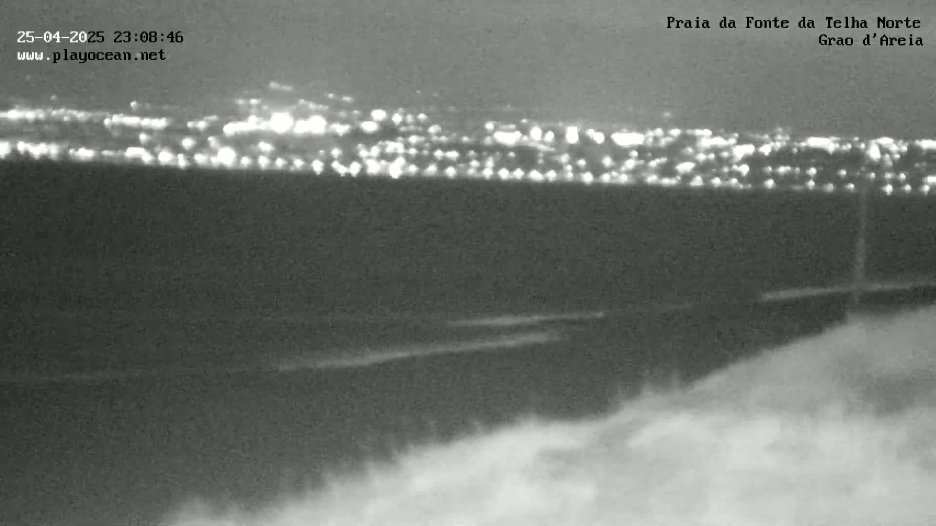 A nighttime, low-light view shows a distant city skyline illuminated by lights, with a foreground of dark, possibly sandy or coastal land under a mostly clear sky.