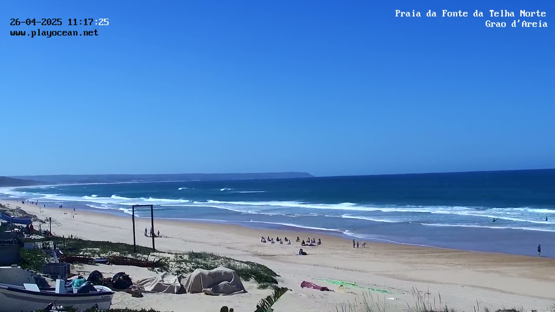A long sandy beach with small waves under a clear blue sky shows several people enjoying the sunny day.
