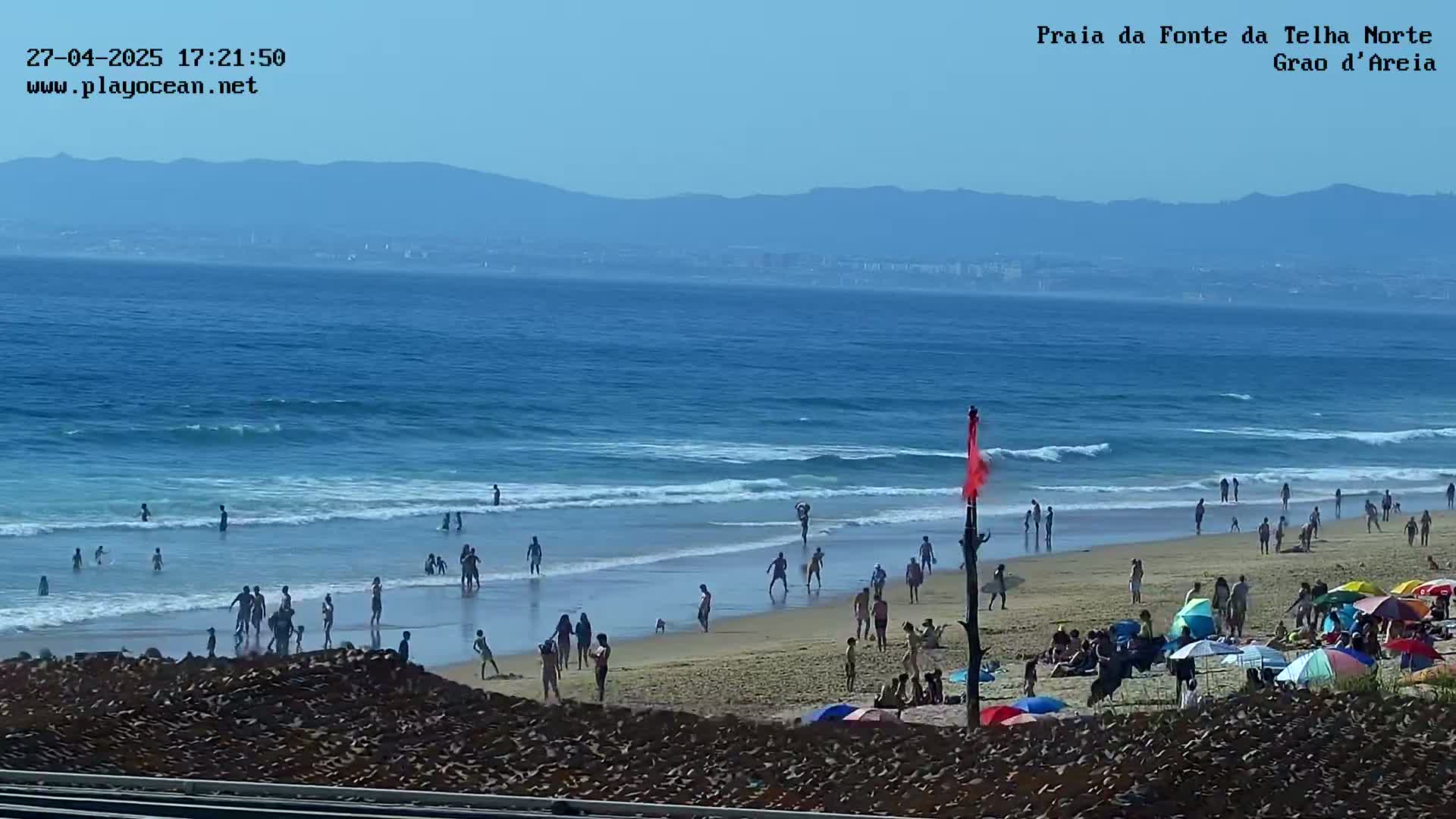 A sandy beach with many people swimming, sunbathing, and relaxing under umbrellas, on a sunny day with blue water and distant mountains.