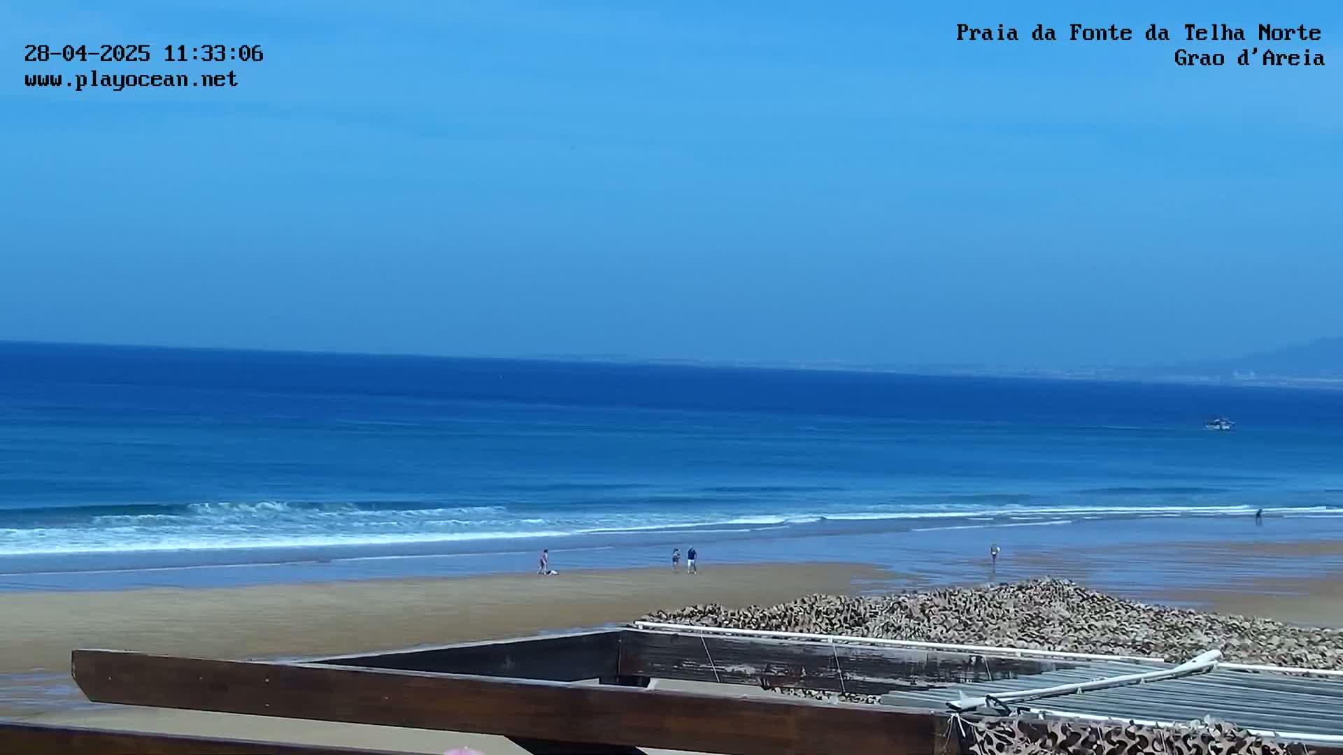 A sandy beach with a few people on it stretches to a calm, blue ocean under a clear sky.