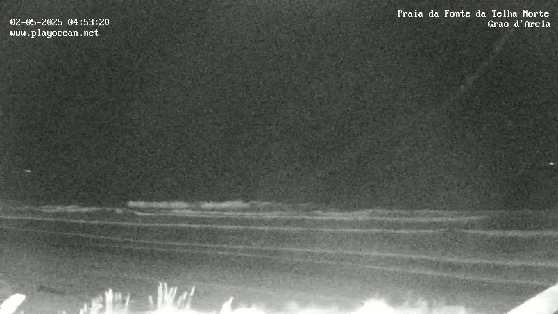 A nighttime, low-light view shows a dark beach with small waves lapping the shore under a cloudy sky.