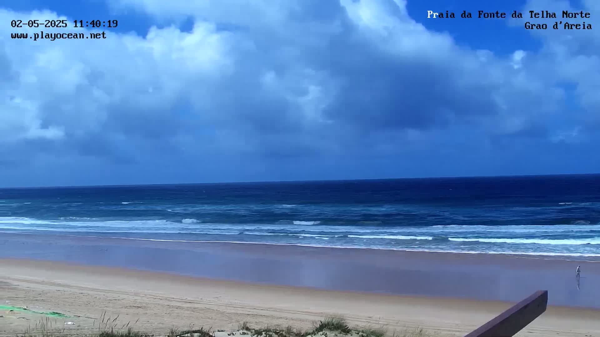 A sandy beach with gently rolling waves under a partly cloudy sky stretches to a dark blue ocean.