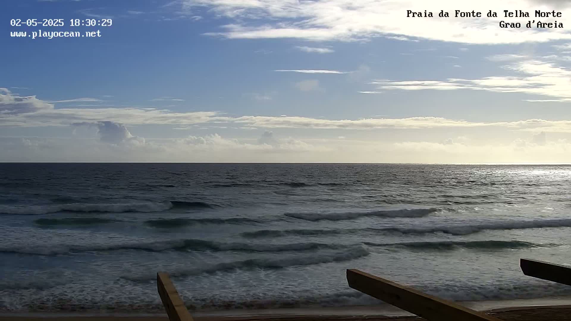 A mostly sunny sky with scattered clouds overlooks a dark blue ocean with small waves breaking on a sandy shore, partially obscured by wooden beams in the foreground.