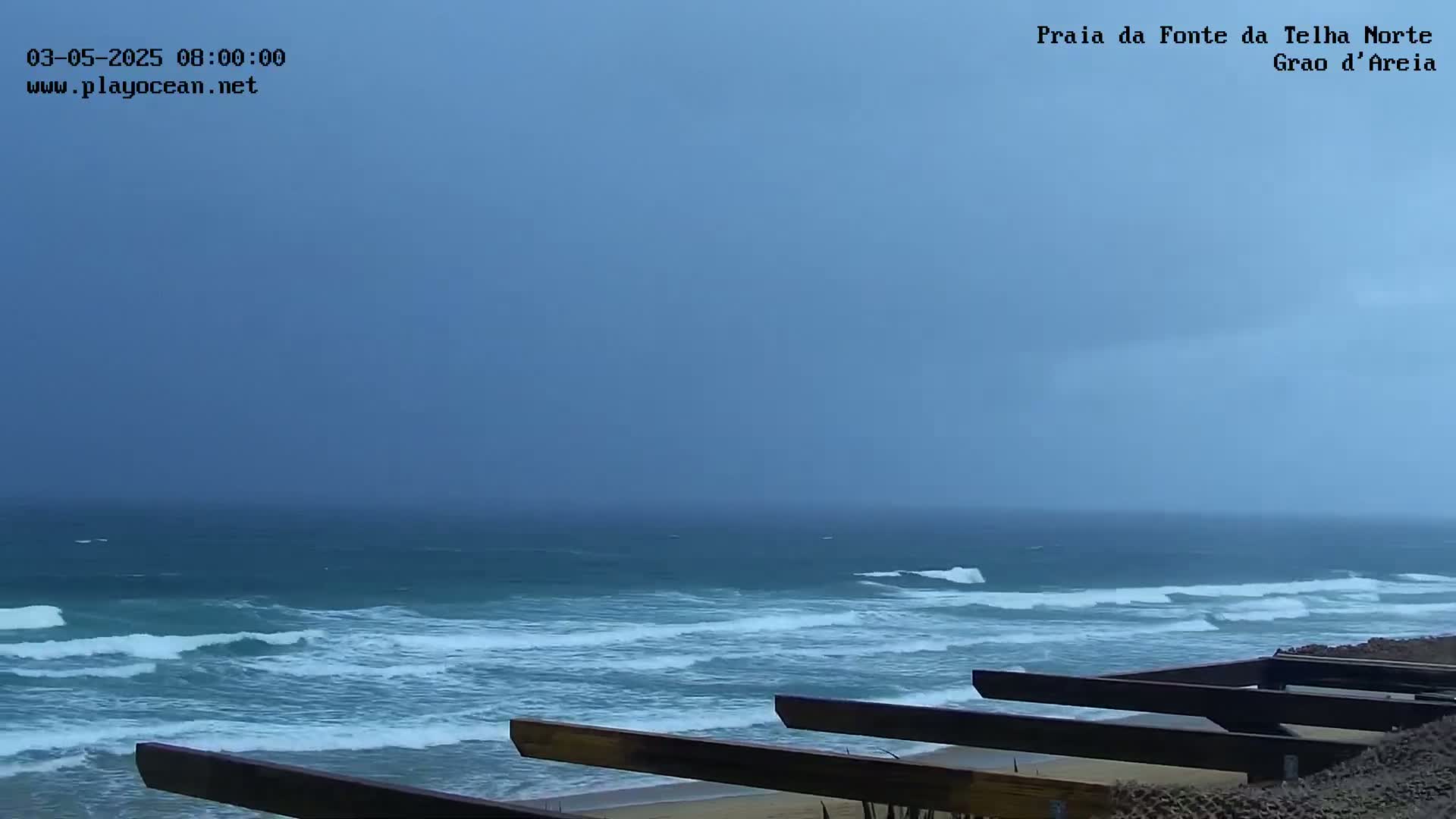A view of a choppy ocean under an overcast sky, seen from behind a low wooden structure on a sandy beach.