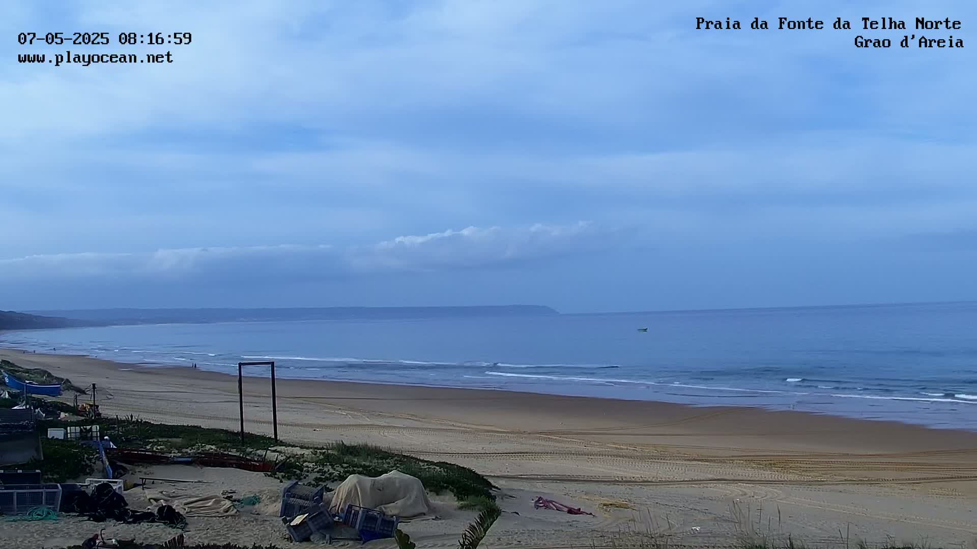 A sandy beach stretches to a calm ocean under a mostly clear, light-blue sky with a few clouds.