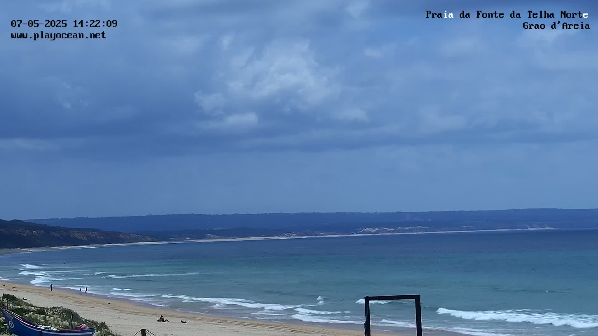 A mostly sunny beach scene shows a wide expanse of light-blue ocean with small waves lapping a sandy shore, backed by a distant coastline under a partly cloudy sky.