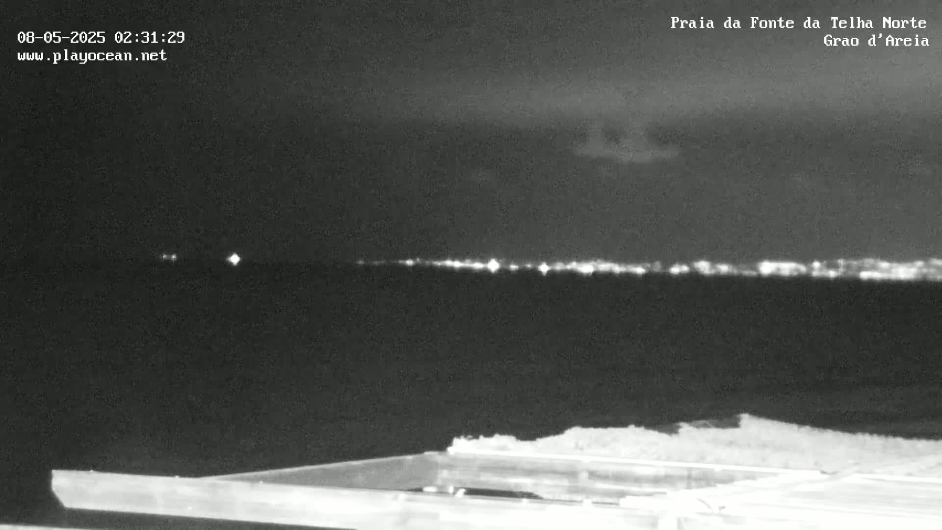 A nighttime, low-light view shows a dark body of water with a distant line of lights on the horizon under a mostly clear sky.