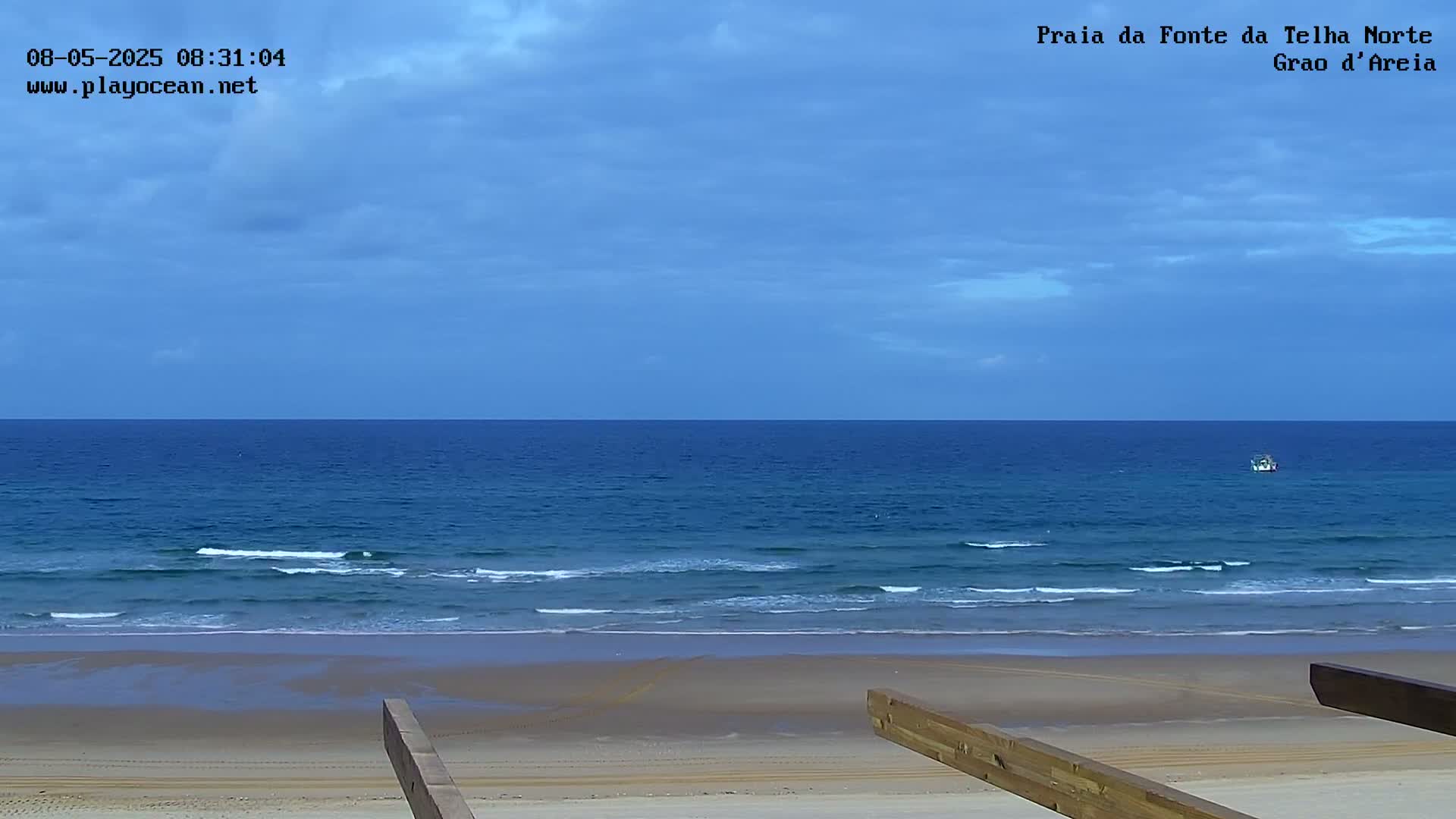 A mostly cloudy sky overlooks a calm ocean with small waves breaking on a sandy beach, viewed from a wooden structure.