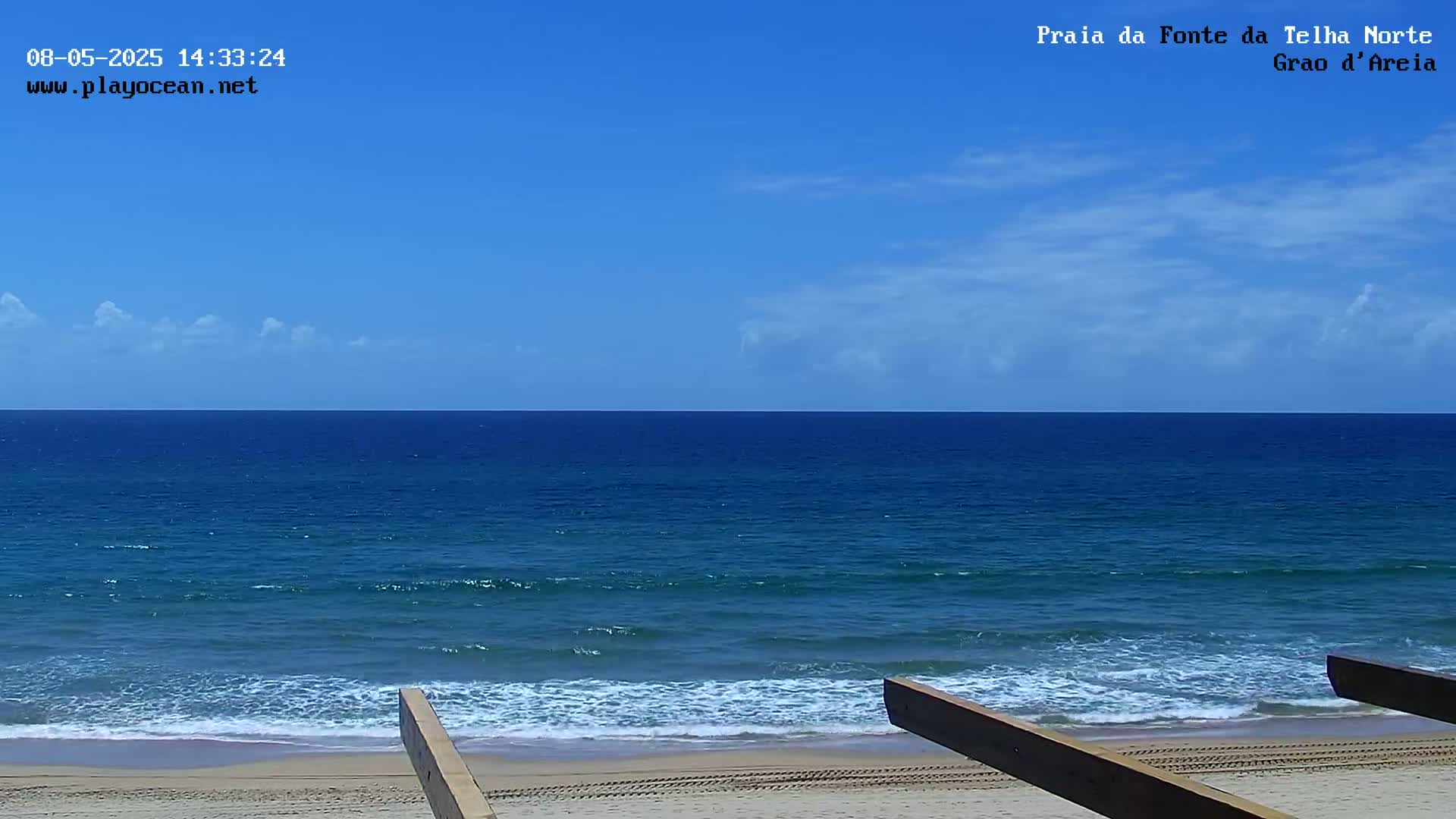 A mostly sunny sky overlooks a tranquil ocean with small waves lapping a sandy beach, partially obscured by wooden structures.