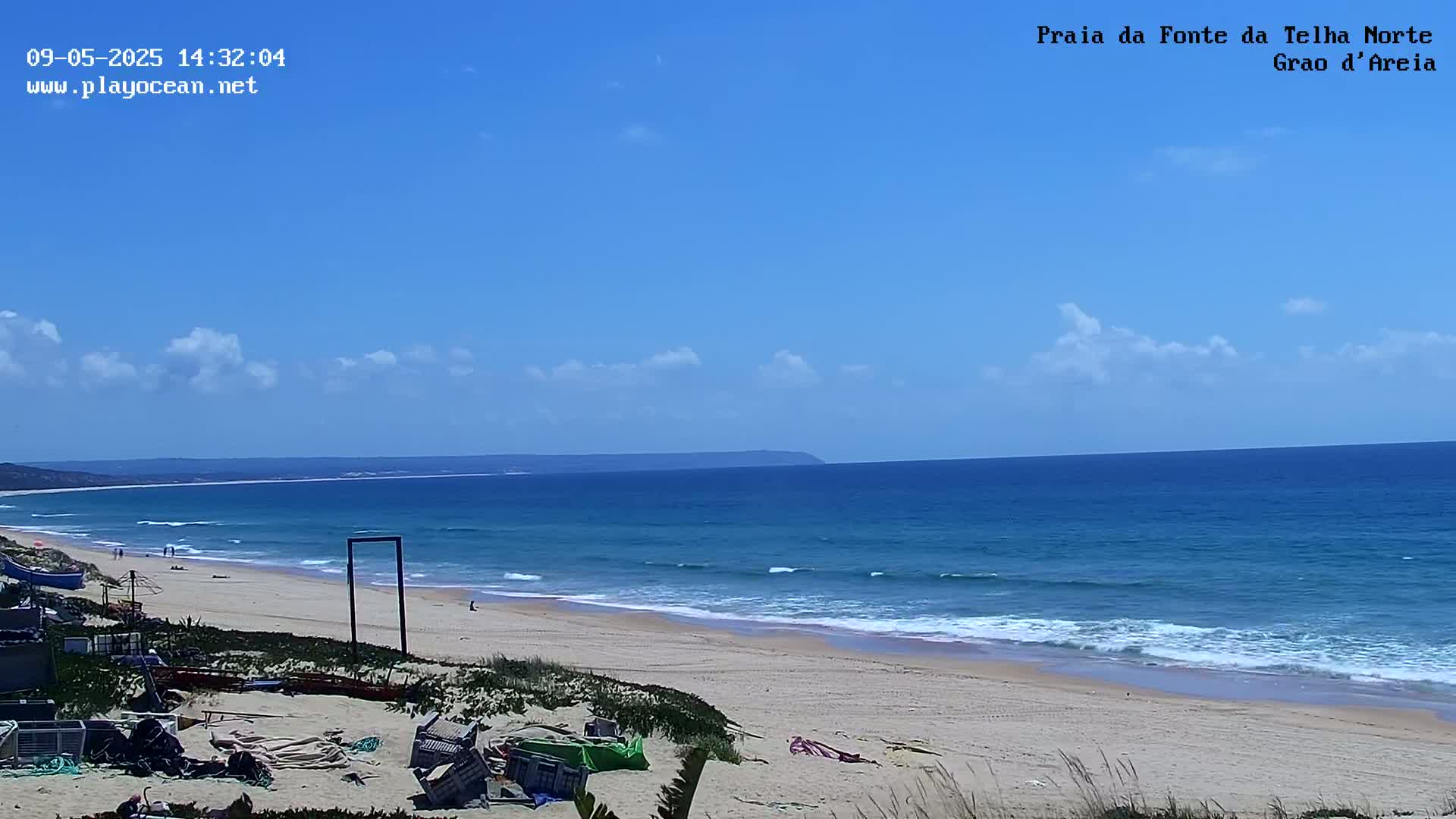 A sandy beach with a calm ocean under a mostly clear blue sky stretches to a distant landmass.