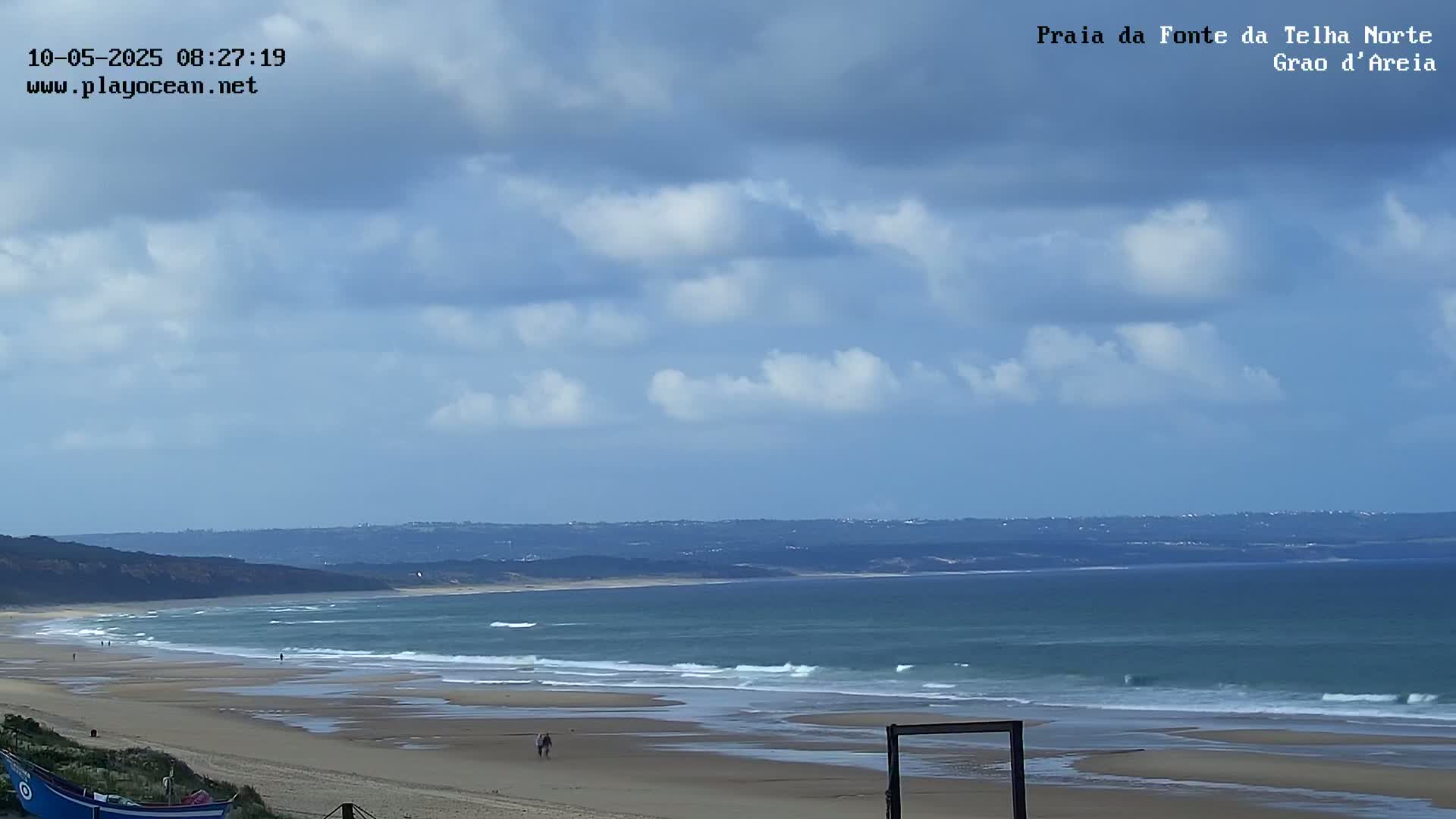 A partly cloudy sky overlooks a long sandy beach with gentle waves rolling onto the shore.
