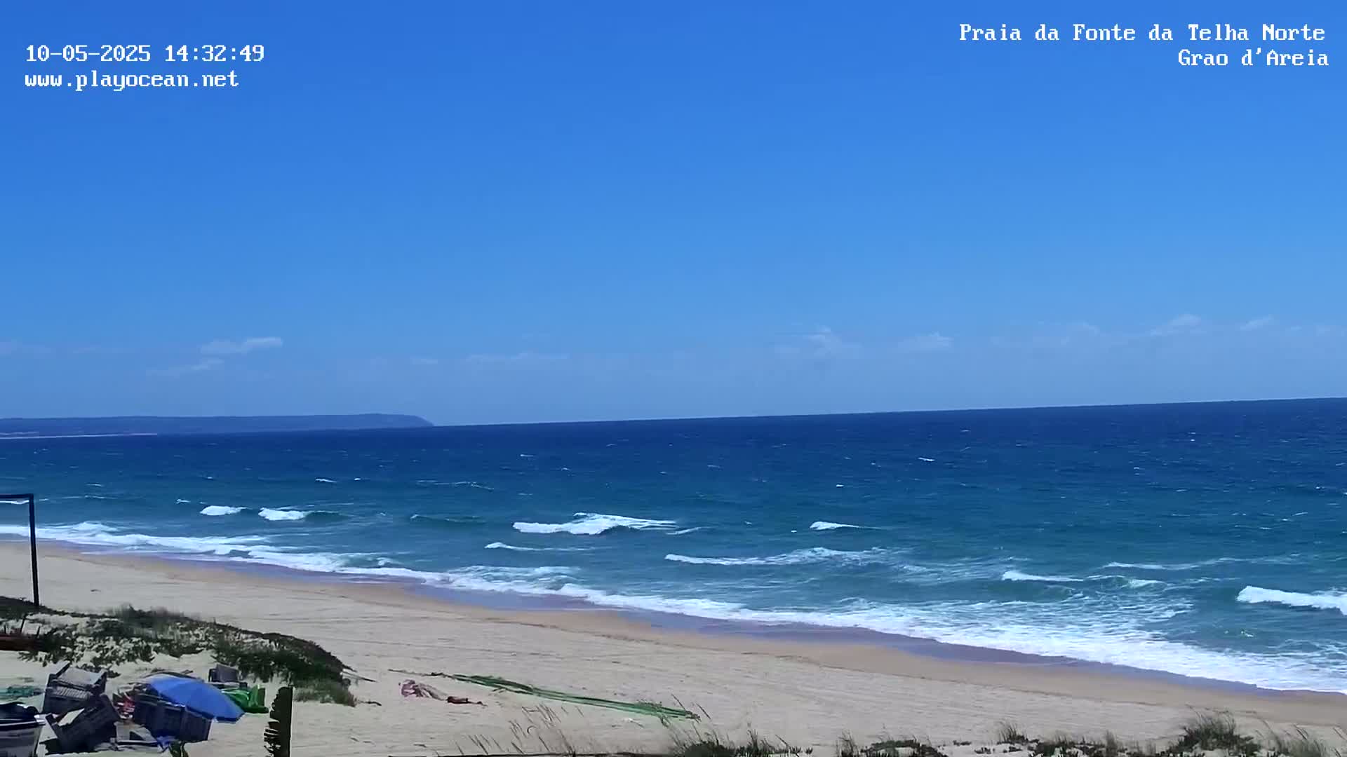 A sandy beach with gently rolling waves under a clear blue sky meets a calm ocean with a distant landmass on the horizon.