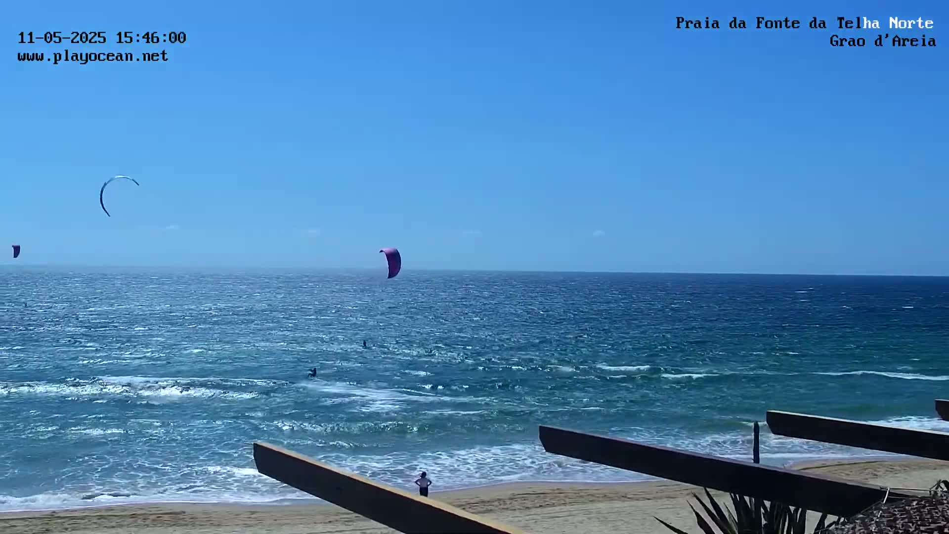 A sandy beach with a few people is seen from behind a wooden structure, overlooking a blue ocean with several kitesurfers on a windy, sunny day.