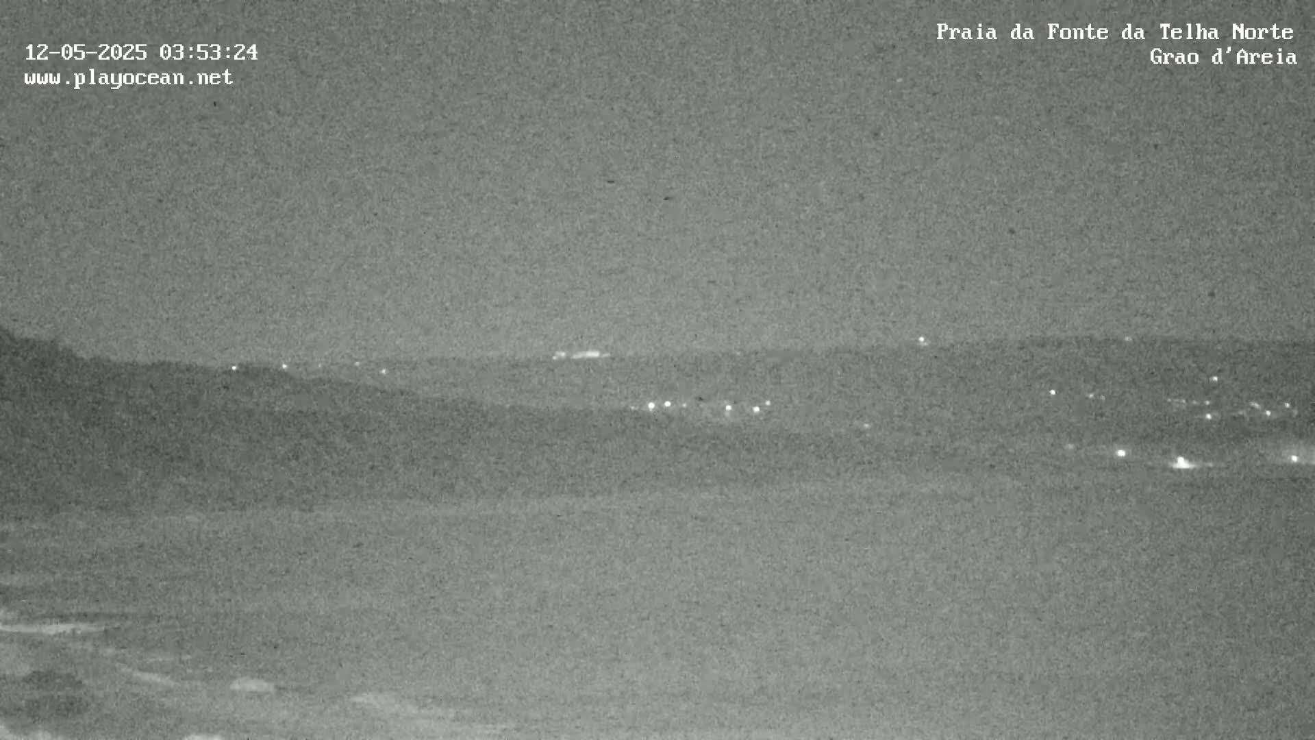 A nighttime, low-light view of a dark coastline with scattered lights visible in the distance.