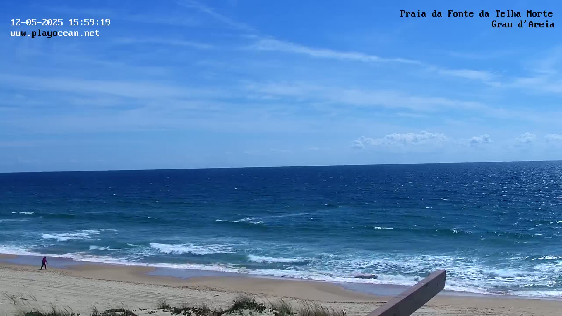 A person walks along a sandy beach with gently rolling waves under a mostly clear blue sky.