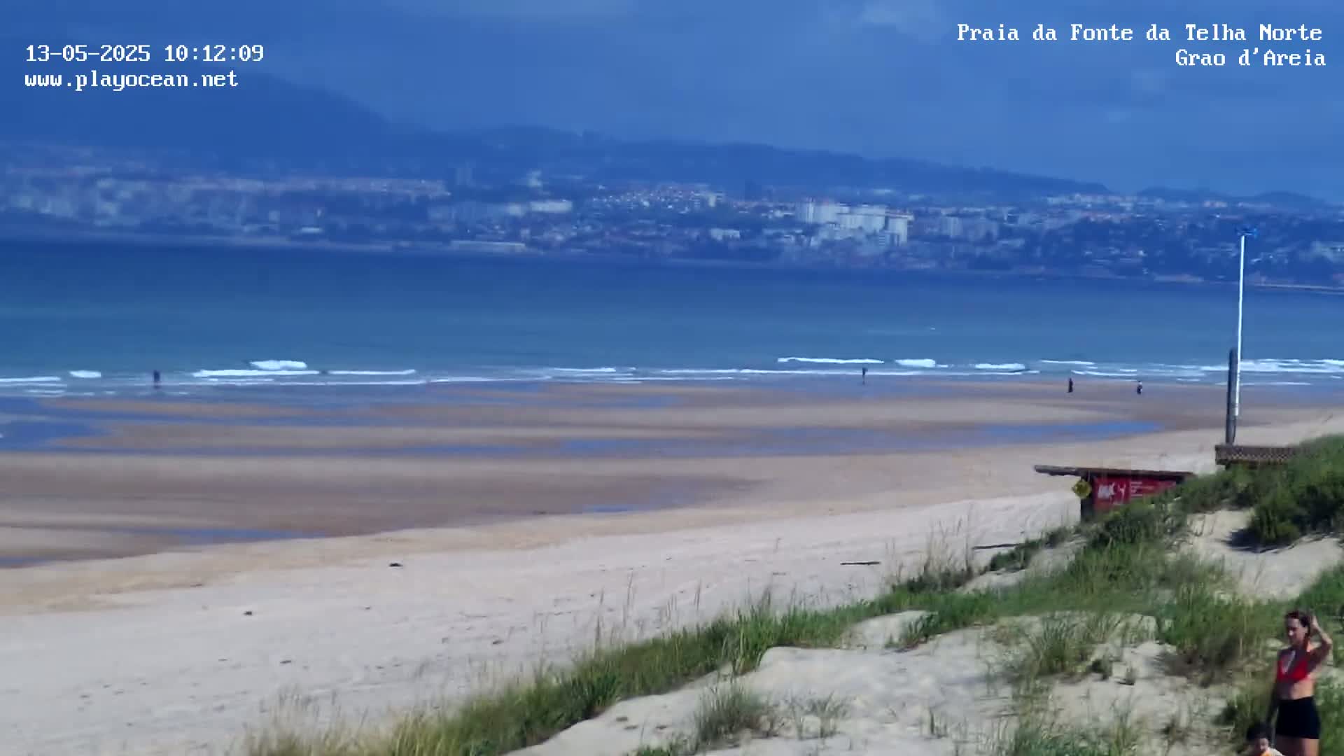 A sandy beach stretches towards a calm ocean under a mostly clear sky, with a distant city visible on the horizon and a few people scattered along the shore.