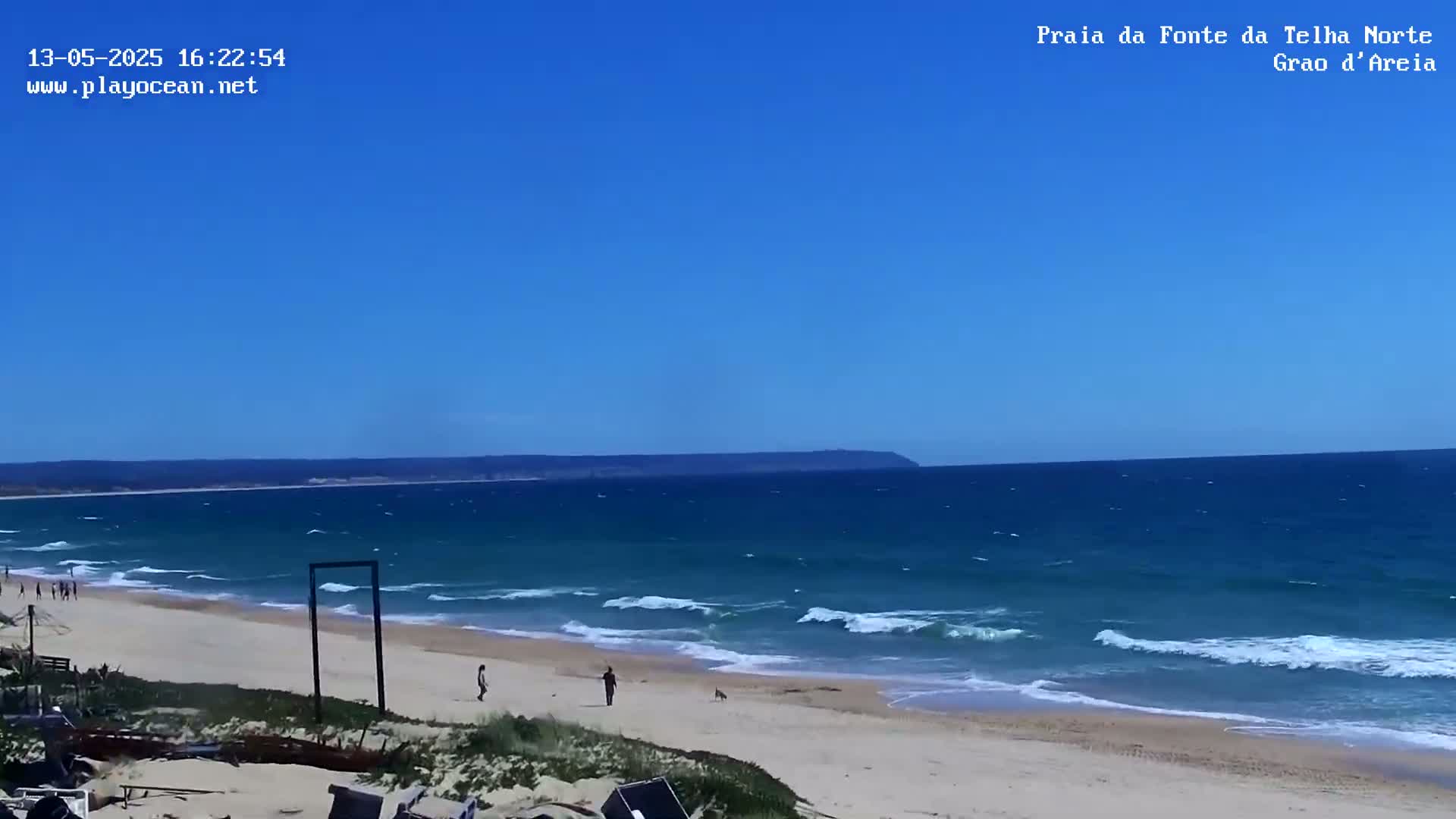A sandy beach with small waves under a clear blue sky shows several people walking along the shore.