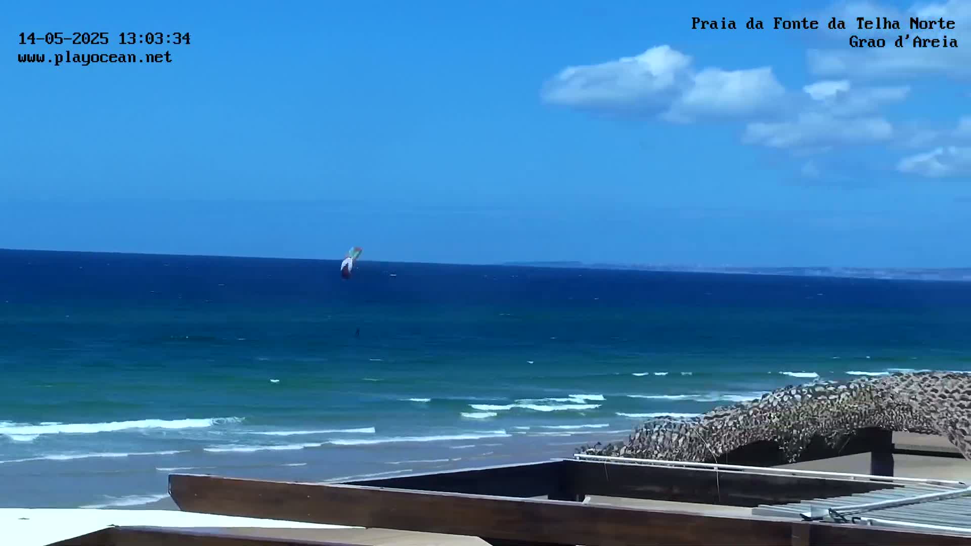 A kitesurfer is visible in the ocean under a mostly sunny sky, with small waves rolling onto a sandy beach.
