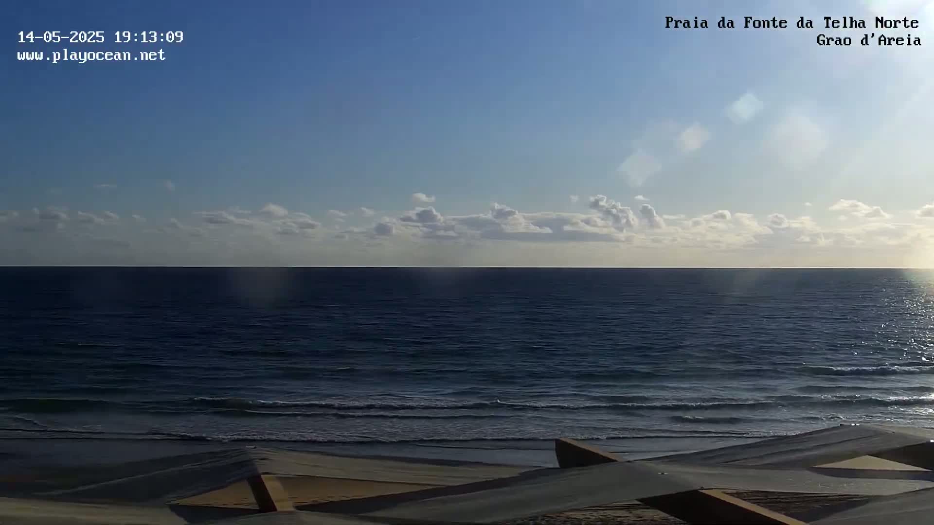 A mostly clear blue sky over a dark blue ocean with small waves lapping a sandy beach, partially obscured by a tan structure in the foreground.