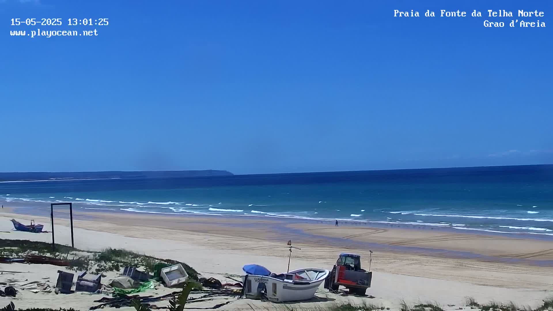 A sandy beach with small waves under a clear blue sky, featuring several small boats and a tractor near the shore.