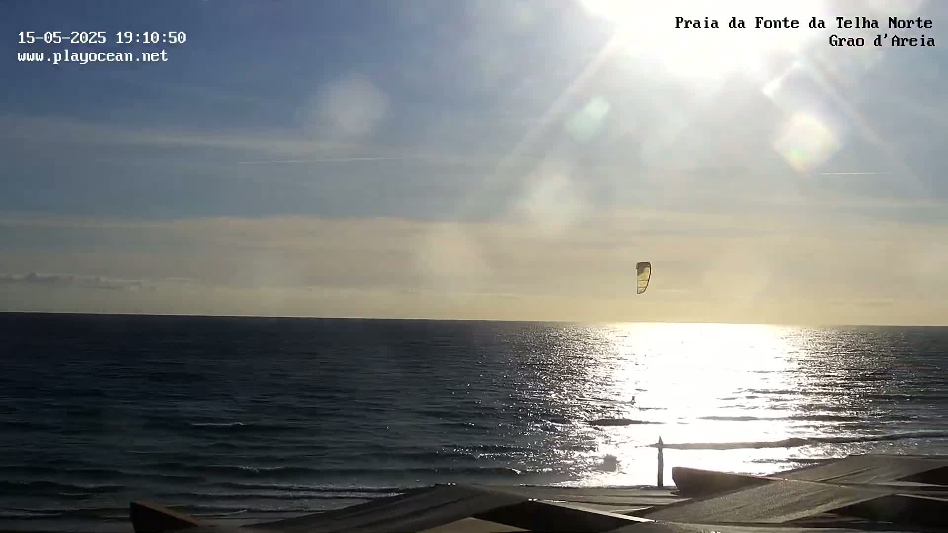 A kitesurfer glides over a bright, sunlit ocean under a mostly clear sky.