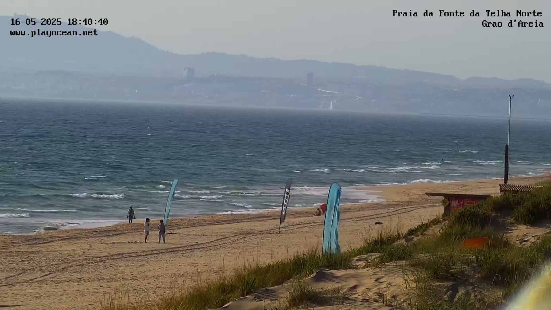 A hazy view shows a sandy beach with a few people and tall flags, gentle waves lapping the shore, and a distant cityscape visible through a light mist.