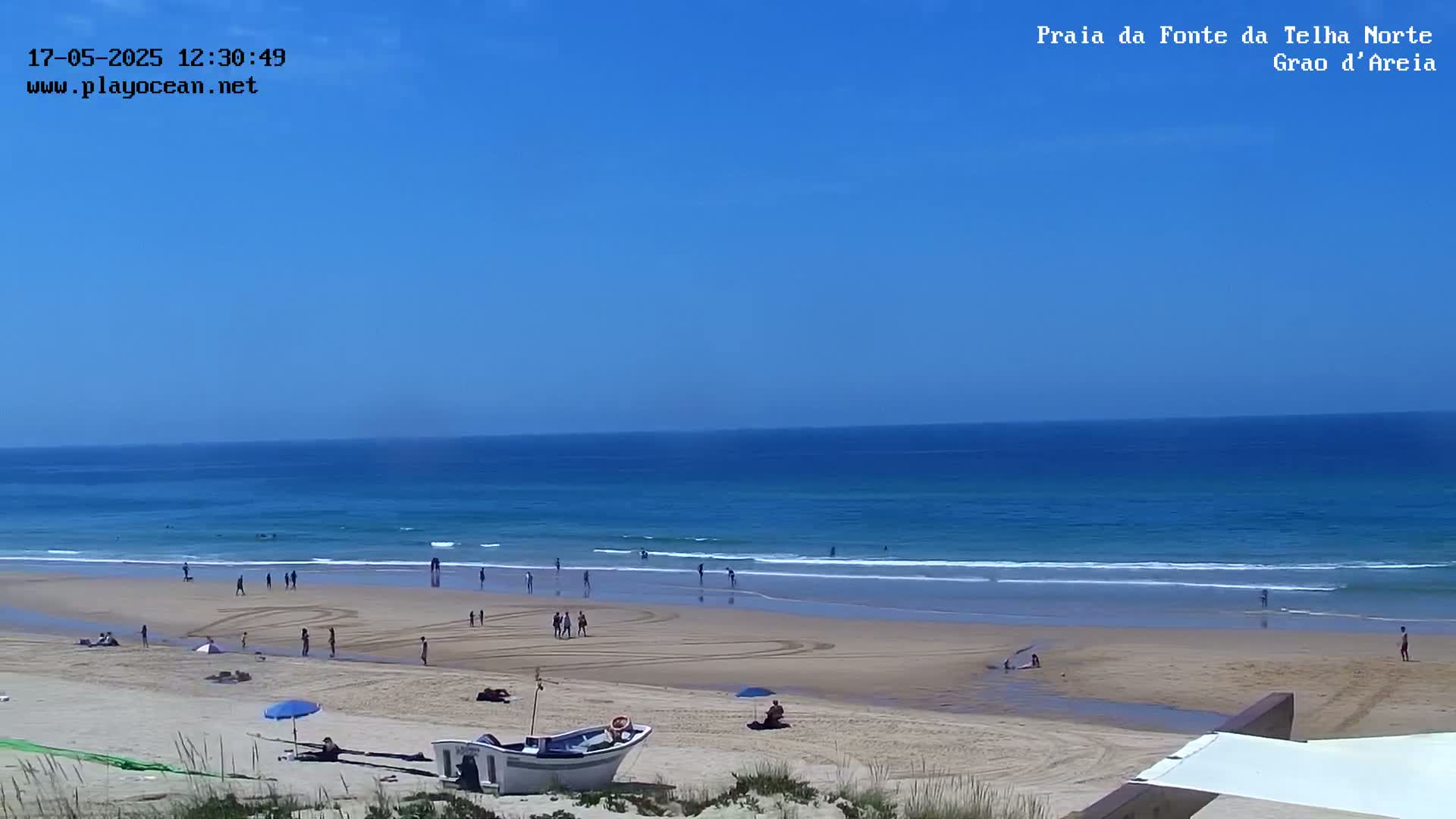 A sandy beach with scattered people, a small boat, and a calm ocean under a clear blue sky.