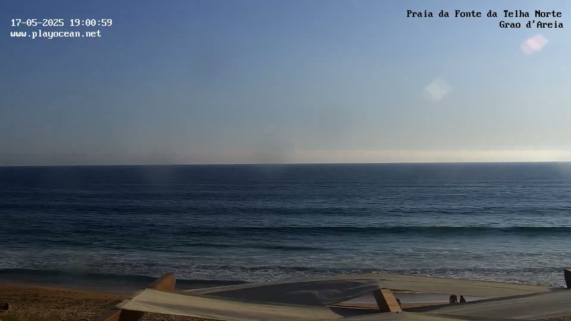 A mostly clear sky overlooks a calm ocean with small waves lapping a sandy beach.