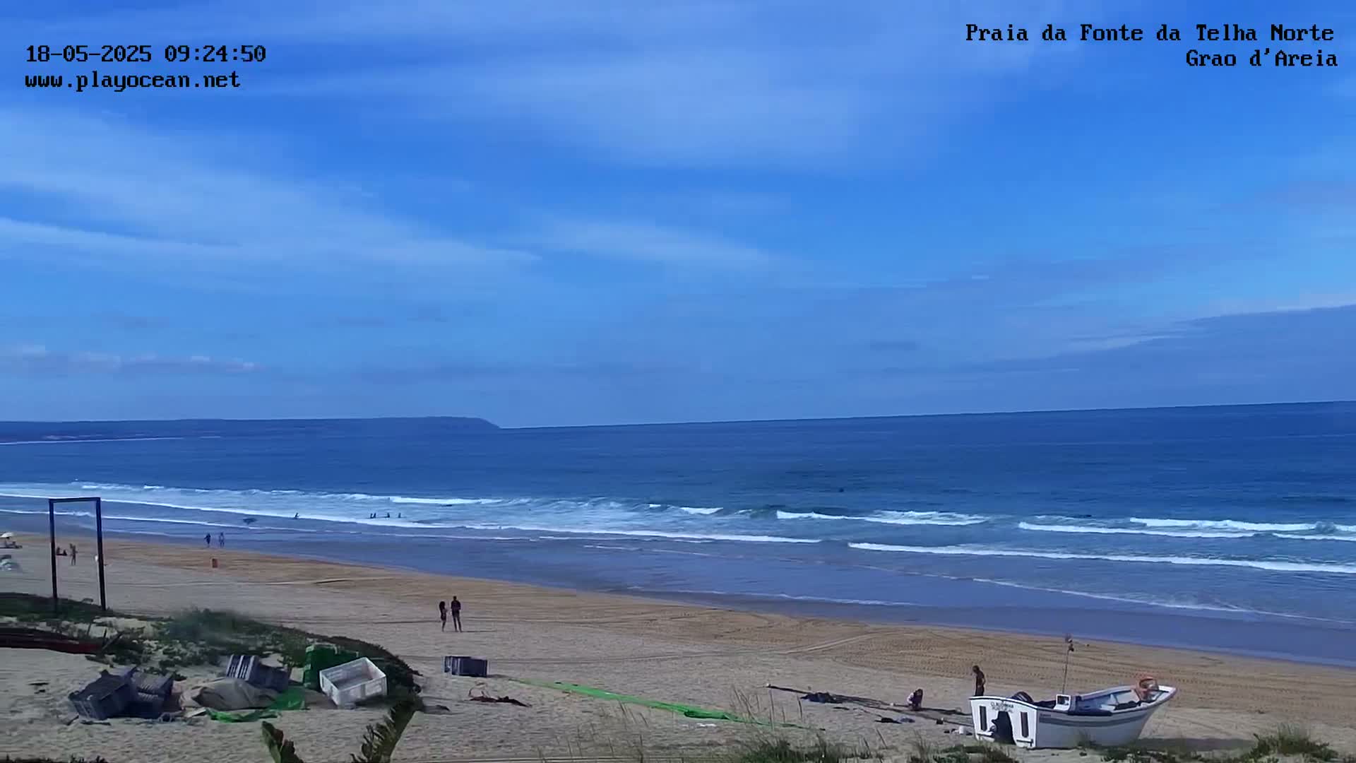 A sandy beach with a few people and a small boat rests on the shore under a mostly clear blue sky, with gentle waves rolling in from a calm ocean.