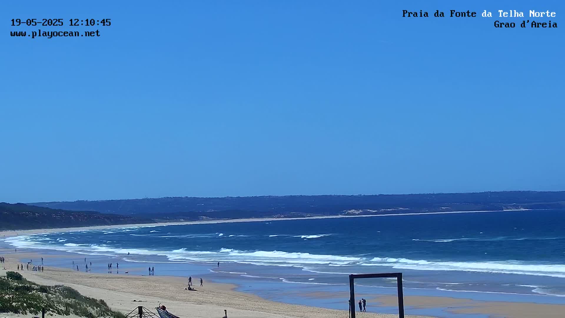 A long sandy beach with gentle waves under a clear blue sky shows people enjoying the sunny day.