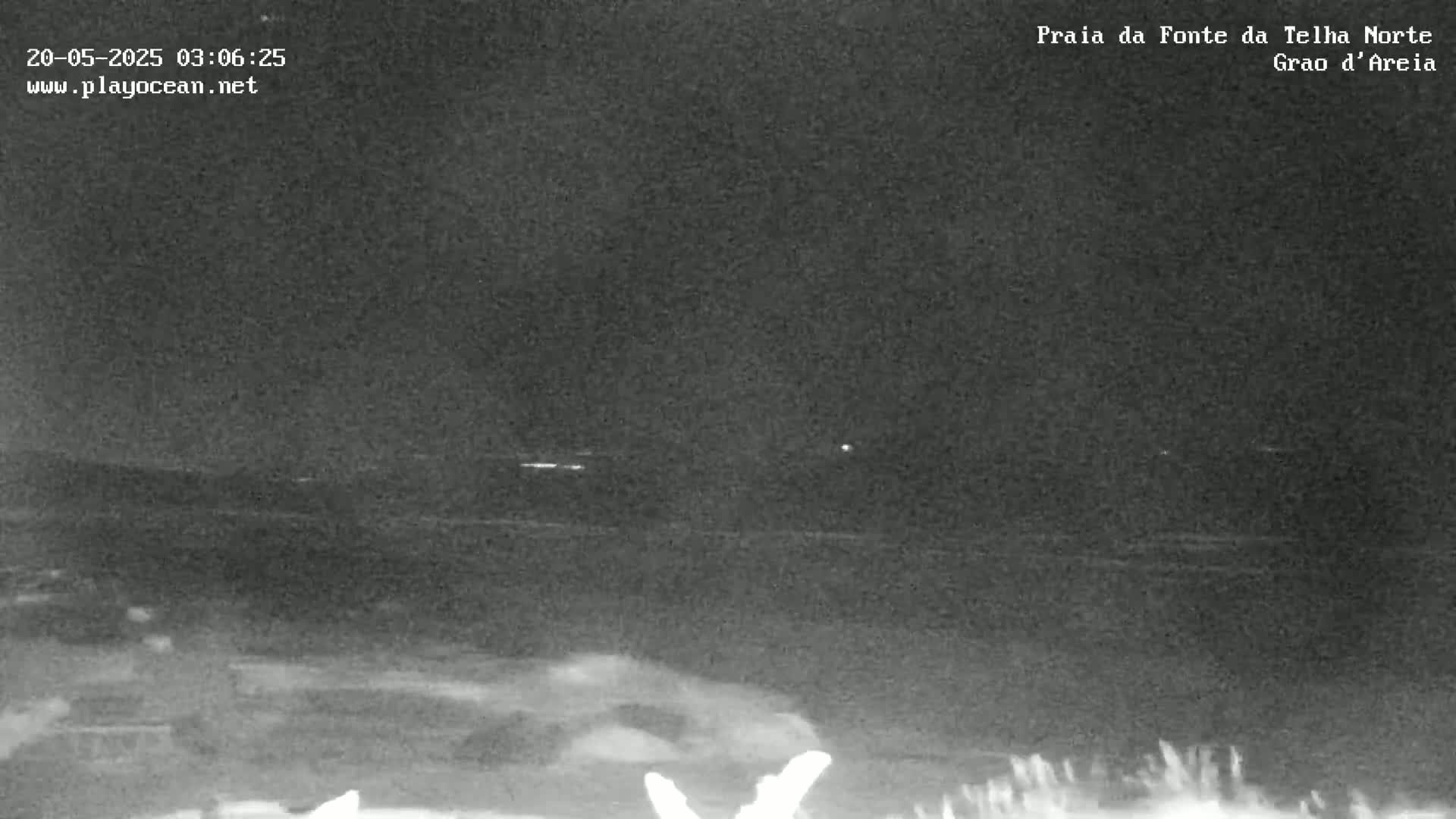 A nighttime, low-light view of a dark ocean shore with faint lights visible in the distance.