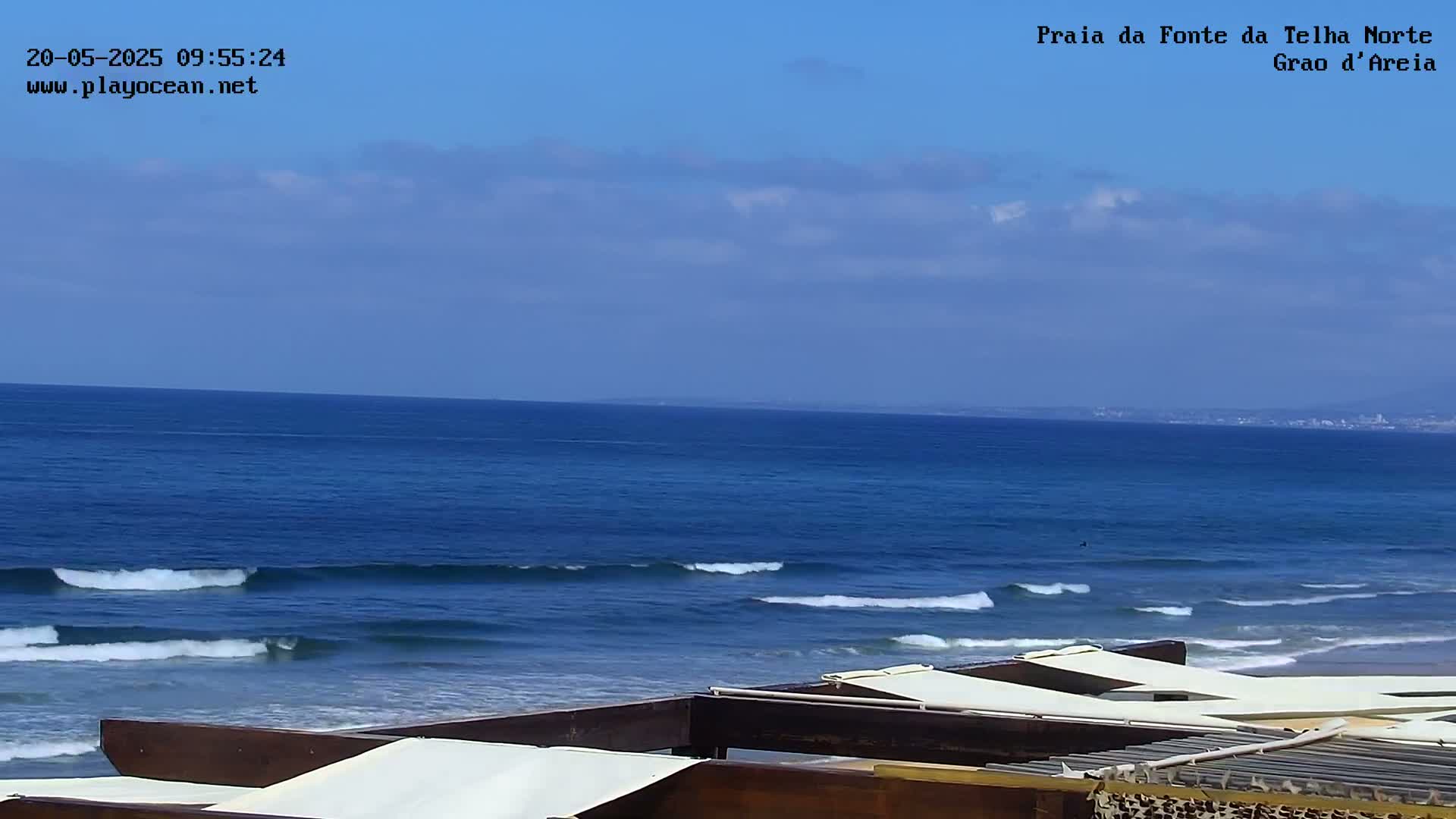 A partly cloudy blue sky overlooks a dark blue ocean with small waves lapping a sandy shore, where several white covered beach loungers are visible.