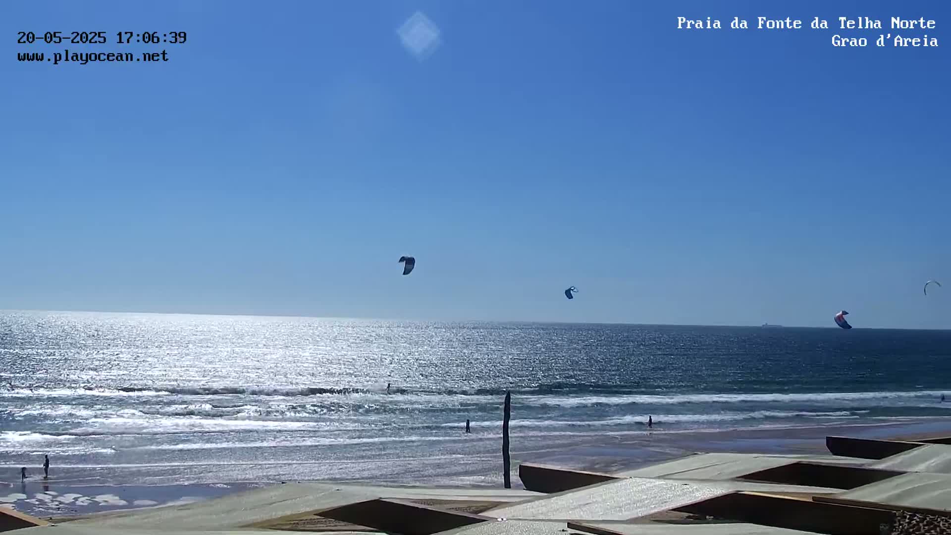 A sunny day at a beach shows several kitesurfers in the ocean, with calm waves and a few people on the sand.
